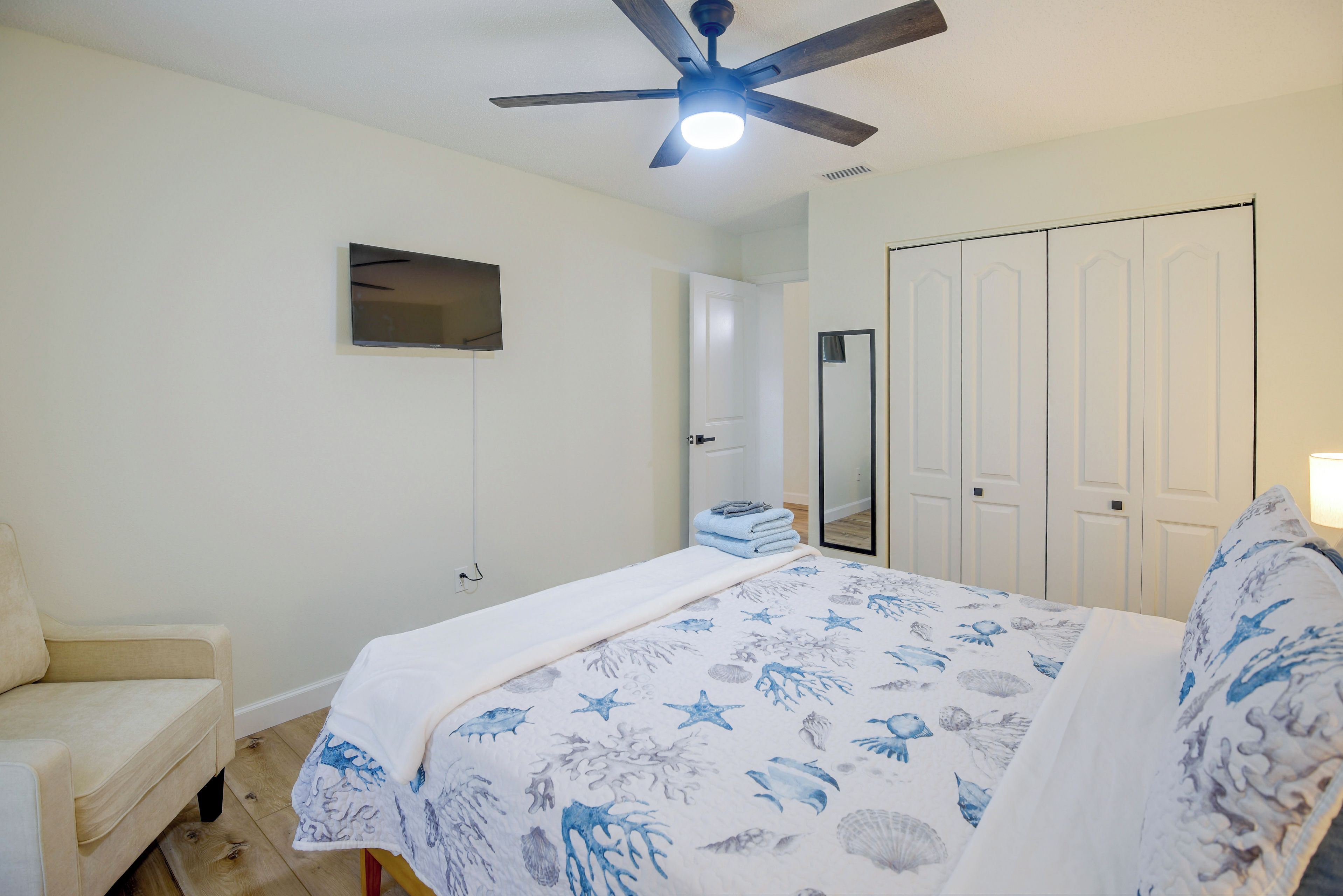 A bedroom with a bed, a ceiling fan, a TV mounted on the wall, a door, and a mirror.