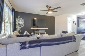 A modern living room with a grey sofa, a flat-screen TV, a ceiling fan, and a large window with blinds.