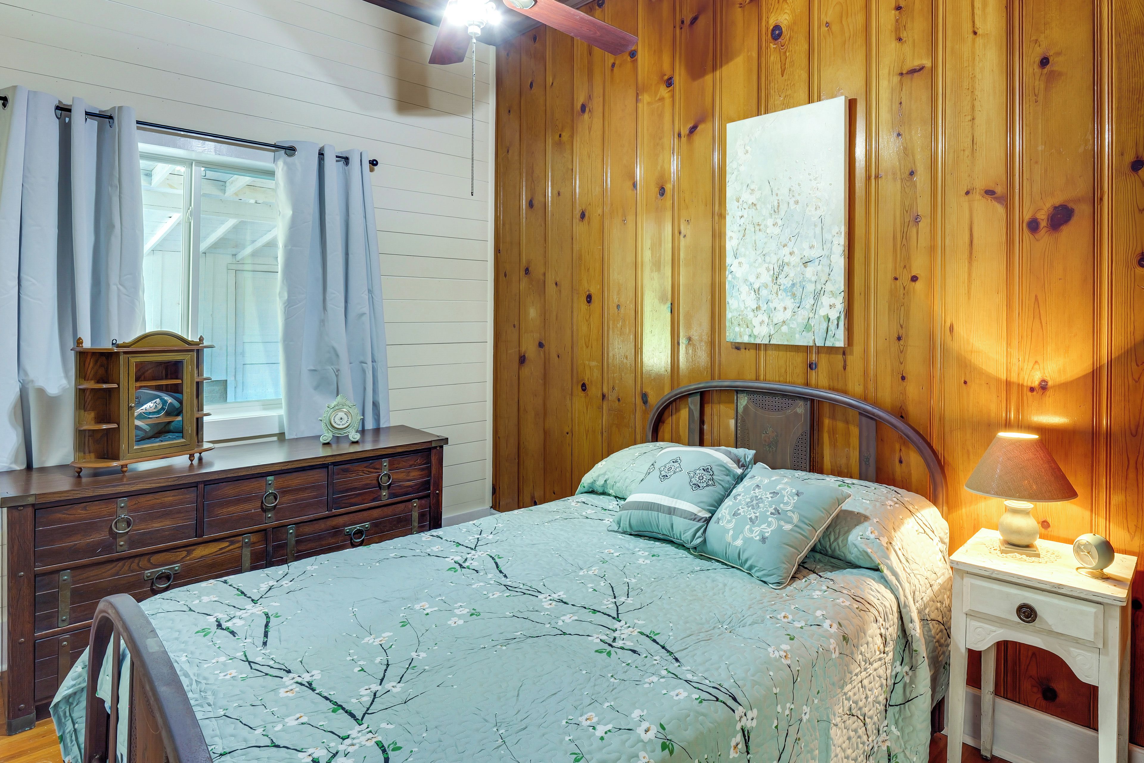 A bedroom with a wooden paneled wall, a bed with floral bedding, a nightstand with a lamp, and a dresser with a mirror.