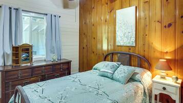 A bedroom with a wooden paneled wall, a bed with floral bedding, a nightstand with a lamp, and a dresser with a mirror.