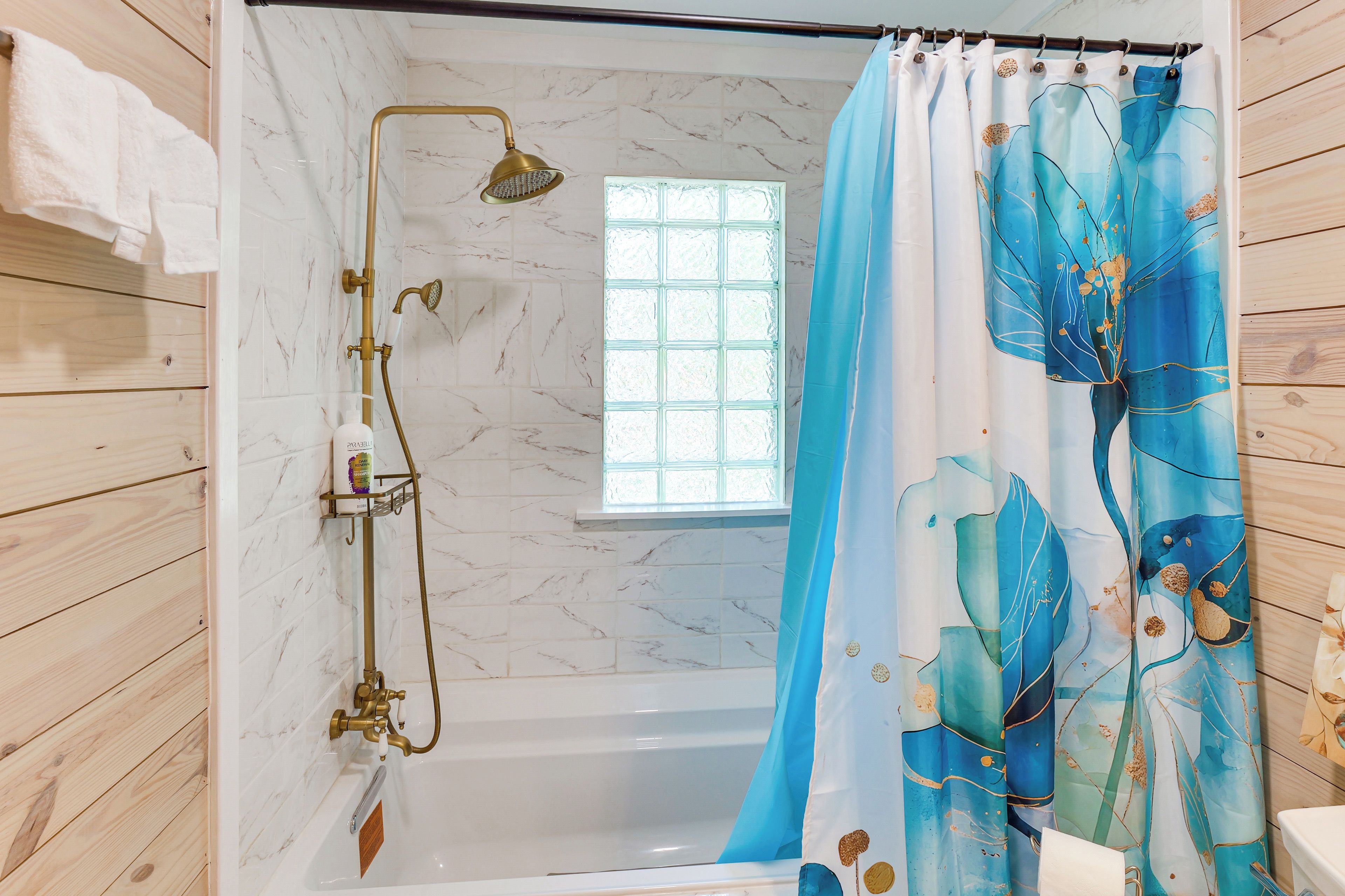 A bathroom with a bathtub, a shower with a gold showerhead, a window, and a curtain with a blue floral pattern.
