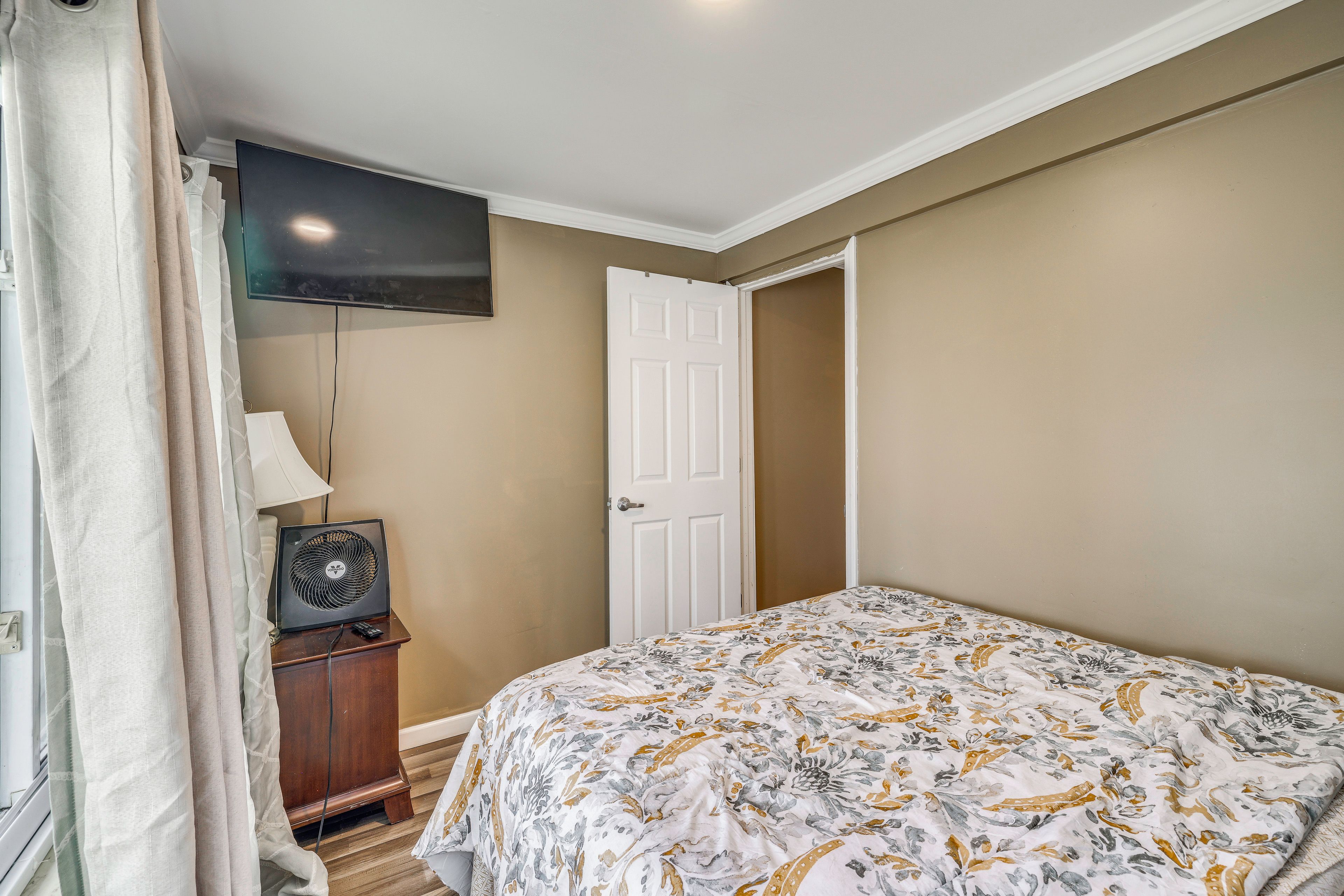 A bedroom with a bed, a television mounted on the wall, a door, and a bedside table with a lamp.