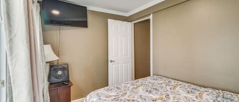A bedroom with a bed, a television mounted on the wall, a door, and a bedside table with a lamp.