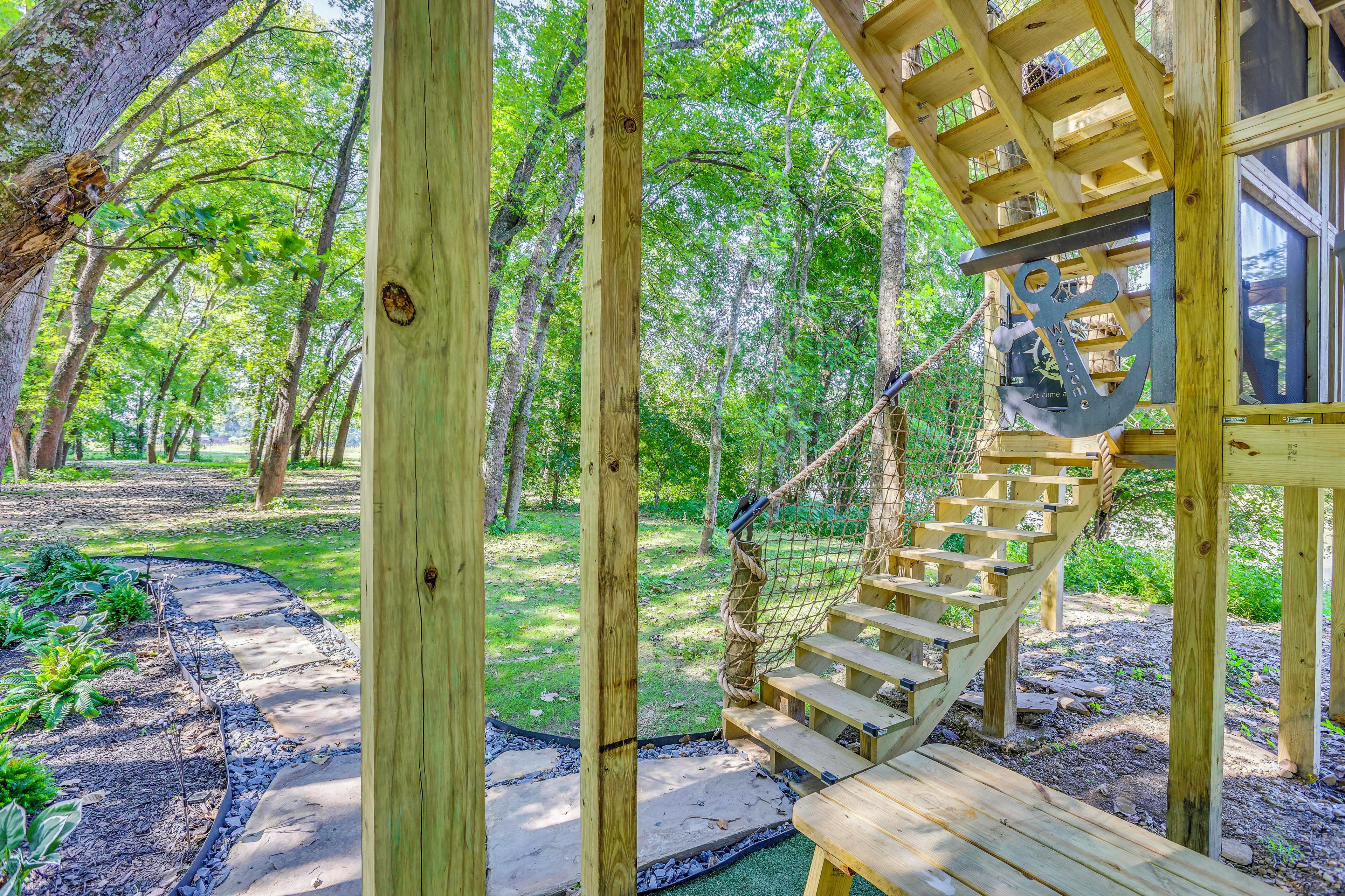 Una escalera de madera conduce a una terraza de madera con un columpio de cuerda, rodeada de árboles y un sendero en el jardín.