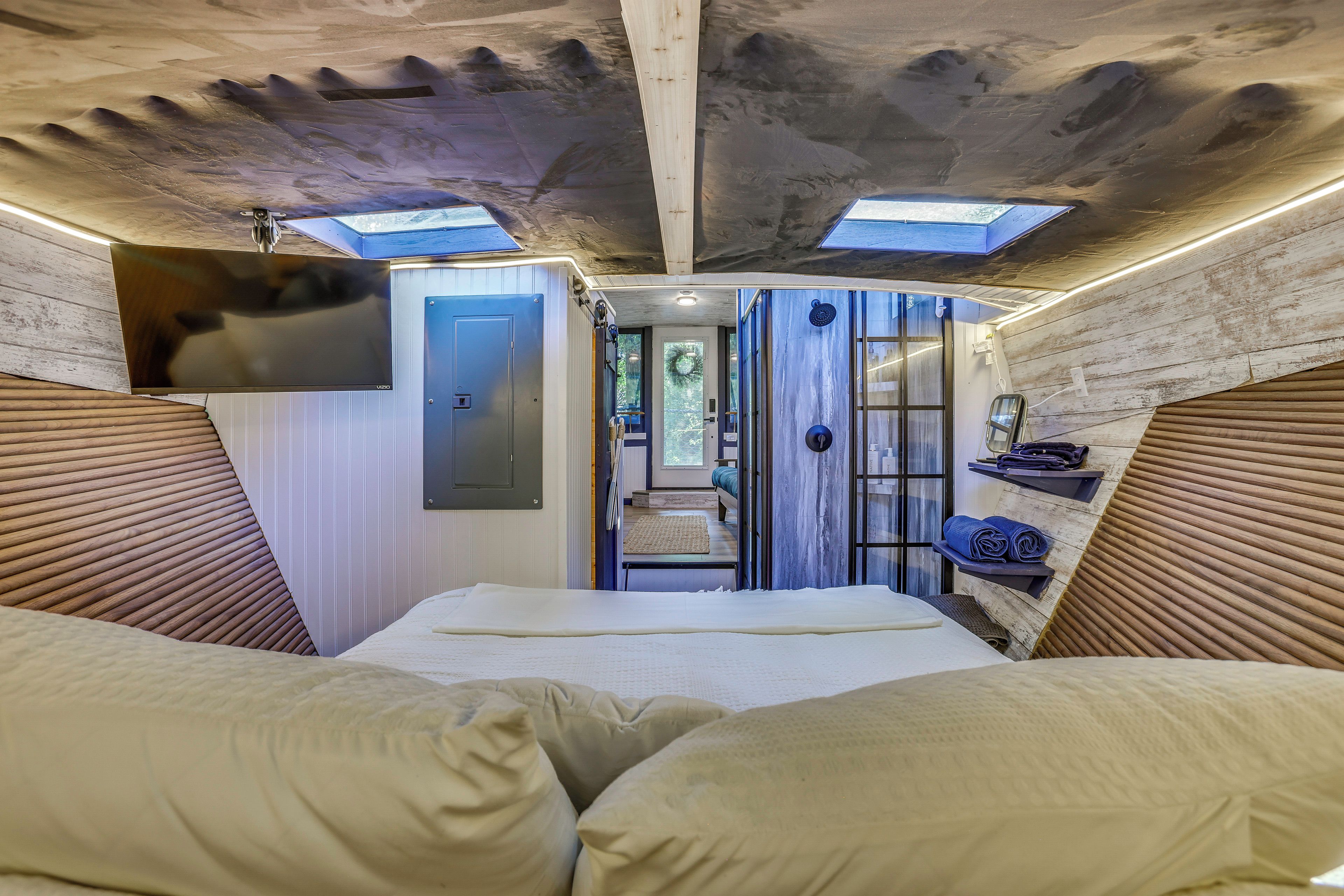 A narrow, elongated room with a bed at the end, a door on the left, and a shower area with a towel rack on the right.