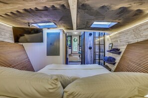 Een lange, smalle kamer met een bed aan het einde, een deur links en een douchegedeelte met een handdoekrek rechts.