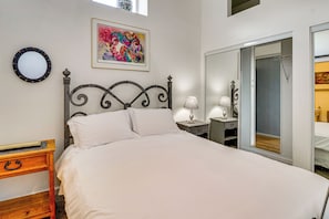 Un dormitorio con una cama, una mesita de noche con lámpara, un aparador, un espejo y un cuadro de colores en la pared.