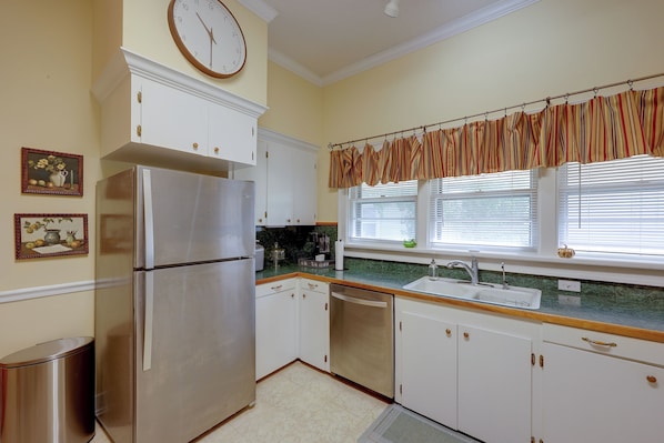 A kitchen with white cabinets, a stainless steel refrigerator, a window with blinds, and a clock on the wall.