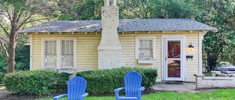 Una casita amarilla con chimenea, dos sillones Adirondack azules adelante y una puerta roja.