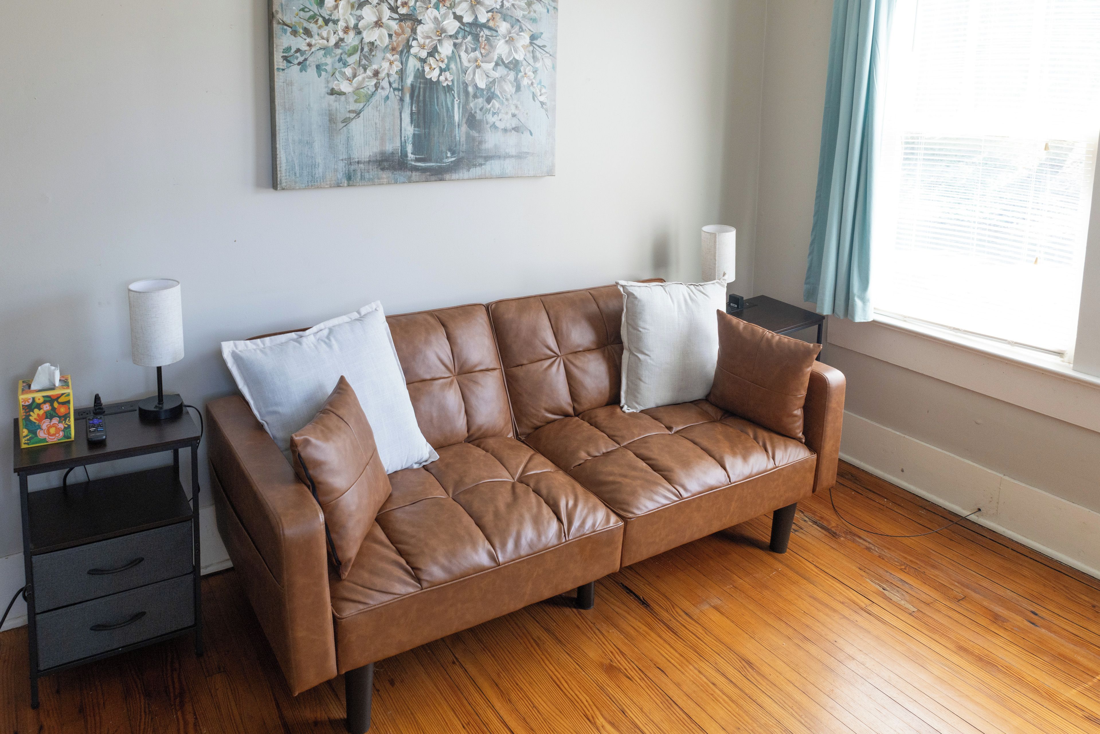 Un sofá de cuero marrón con almohadones blancos en una habitación con piso de madera, una mesita con una lámpara y un cuadro en la pared.