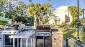 Ein Grillbereich im Freien mit zwei Gasgrills, Picknicktischen und Sonnenschirmen.