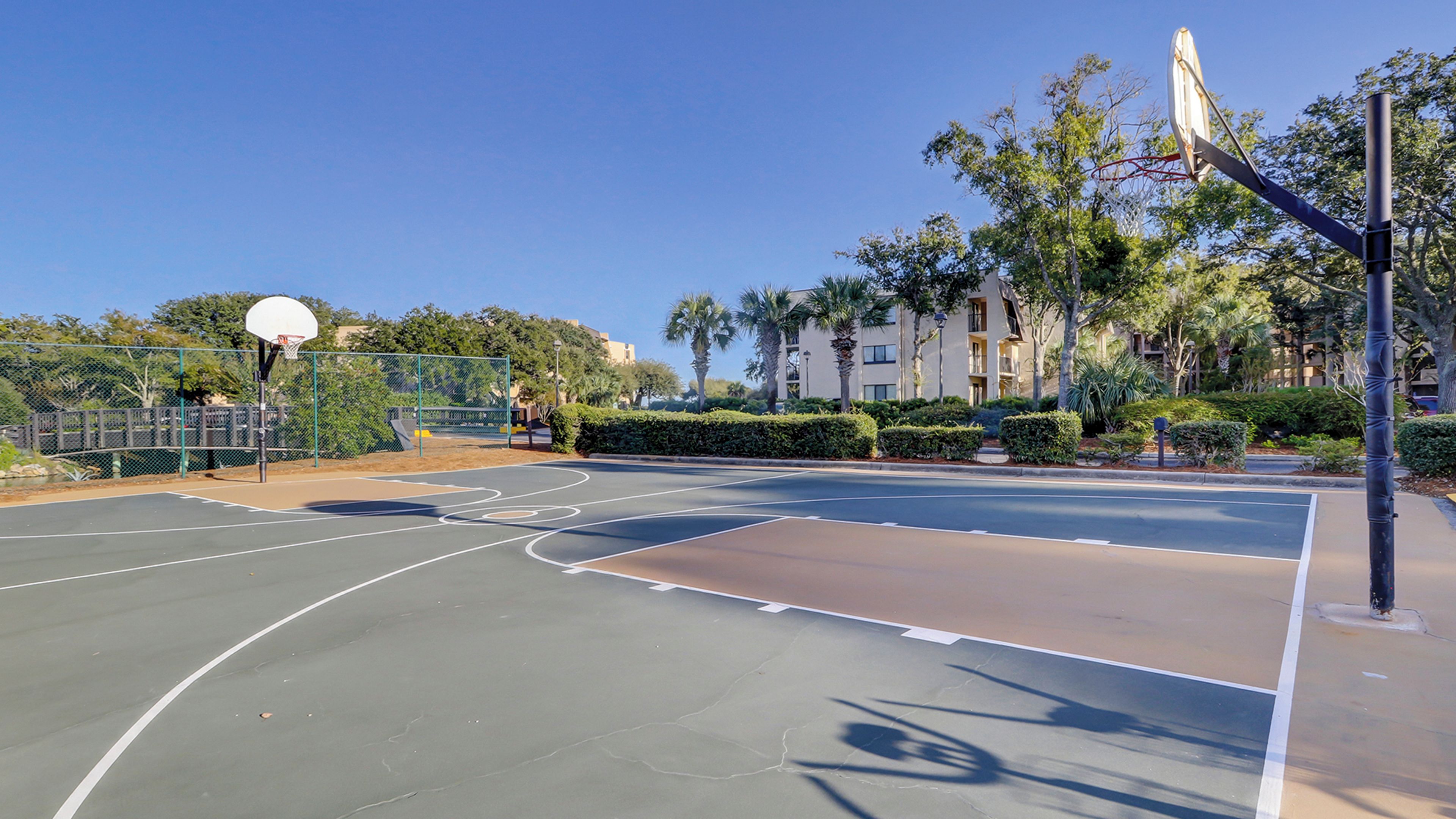 An outdoor basketball court with a basketball hoop, surrounded by a fence and palm trees.