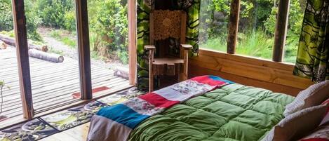 A room with a bed, a chair, and a view of a forest through large windows.