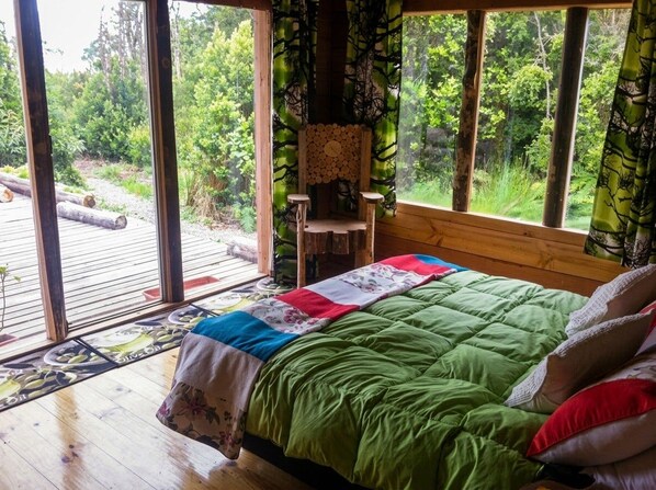 A room with a bed, a chair, and a view of a forest through large windows.