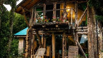 A rustic log cabin with a balcony, surrounded by lush greenery and flowering plants.