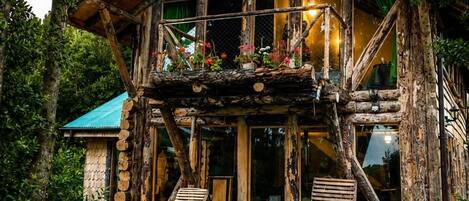 Un chalet rustique en rondins de bois, doté d’un balcon, entouré d’une végétation luxuriante et de plantes en fleurs.