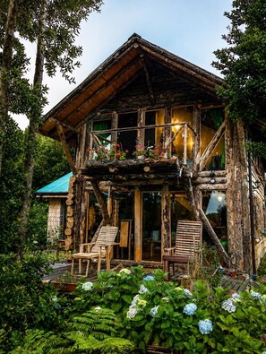 Una rustica baita di legno con un balcone, immersa nel verde e circondata da piante fiorite.