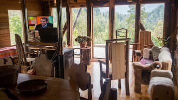A rustic interior with wooden furniture, a television, and large windows offering a forest view.