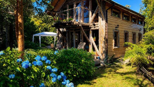 Une maison en bois au style rustique, dotée d’un balcon et entourée d’une végétation luxuriante et de fleurs d’hortensias bleues.