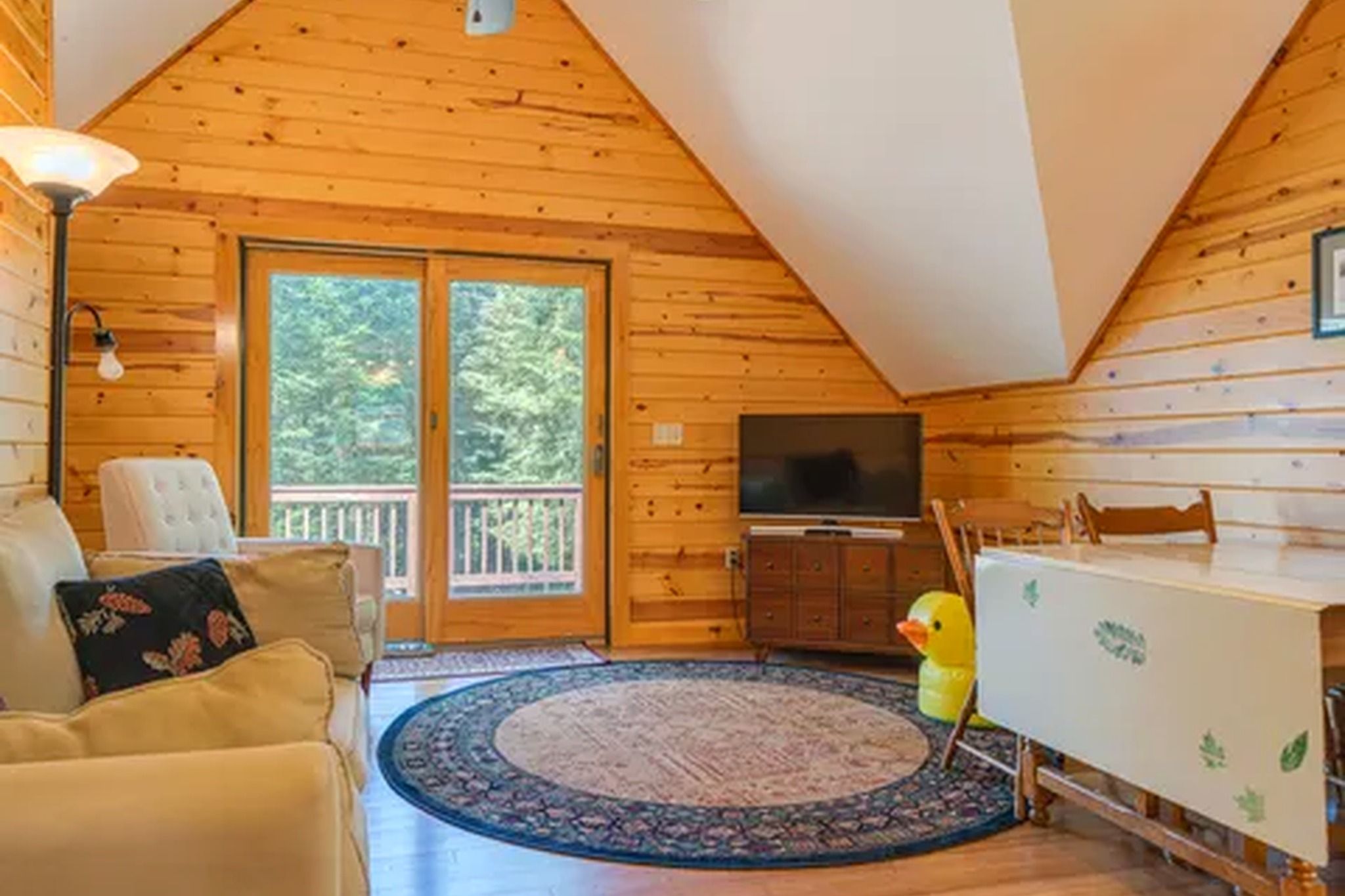 A log cabin interior with a sofa, a television, a dining table, and a window with a view of trees.