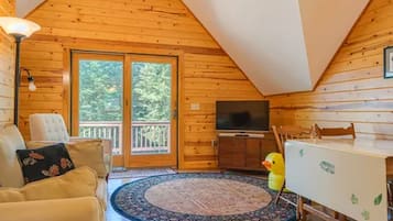A log cabin interior with a sofa, a television, a dining table, and a window with a view of trees.