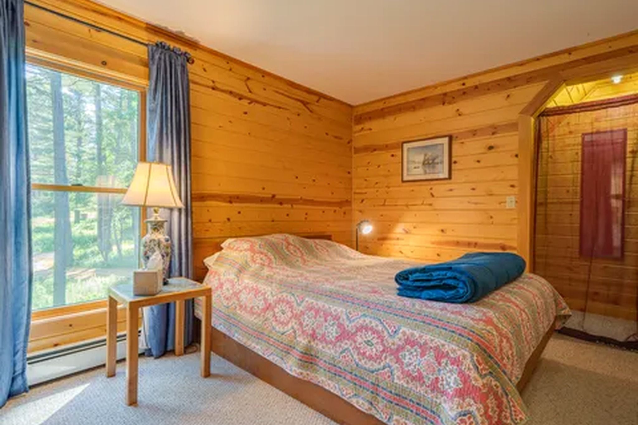 Ein Zimmer aus Holz mit einem Bett, einem Nachttisch, einer Lampe und einem Fenster mit Vorhängen.
