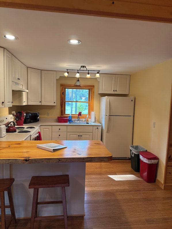 A kitchen with white cabinets, a wooden countertop, a refrigerator, and a window with a view of trees.