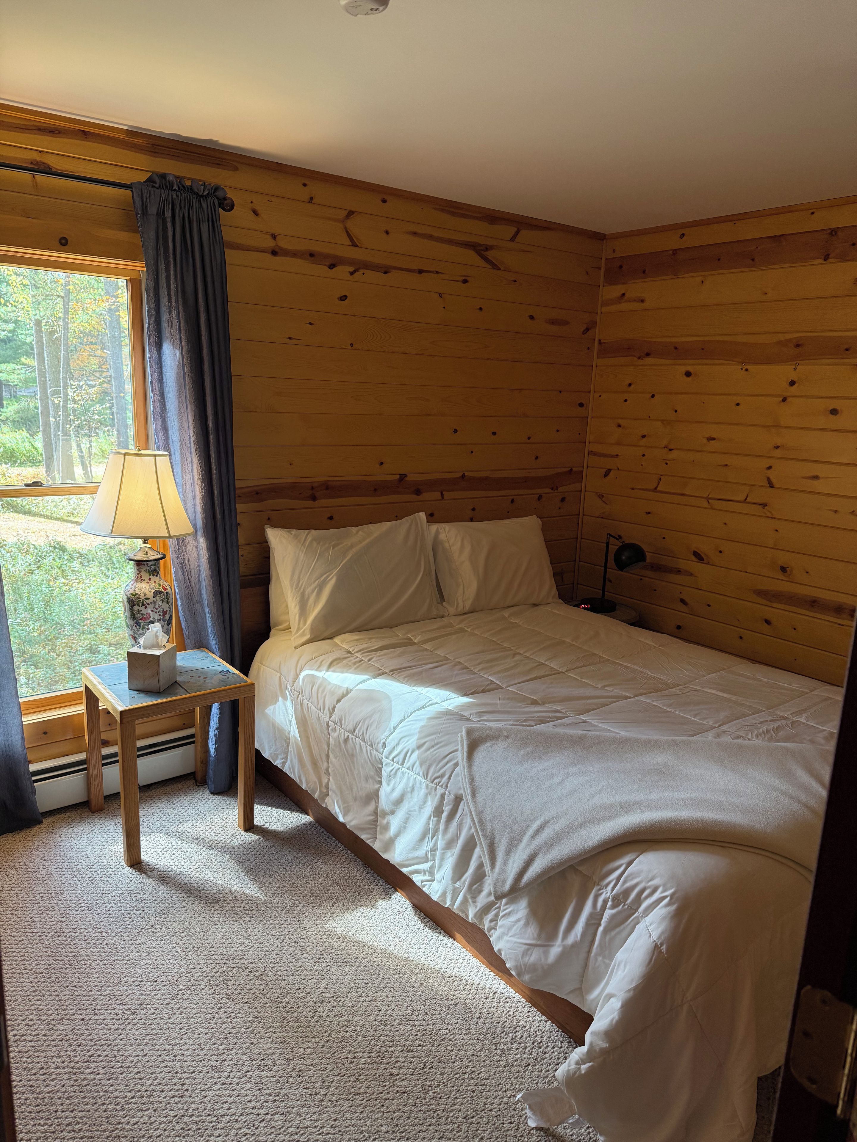 A bedroom with a wooden paneled wall, a bed with white bedding, a bedside table with a lamp, and a window with curtains.