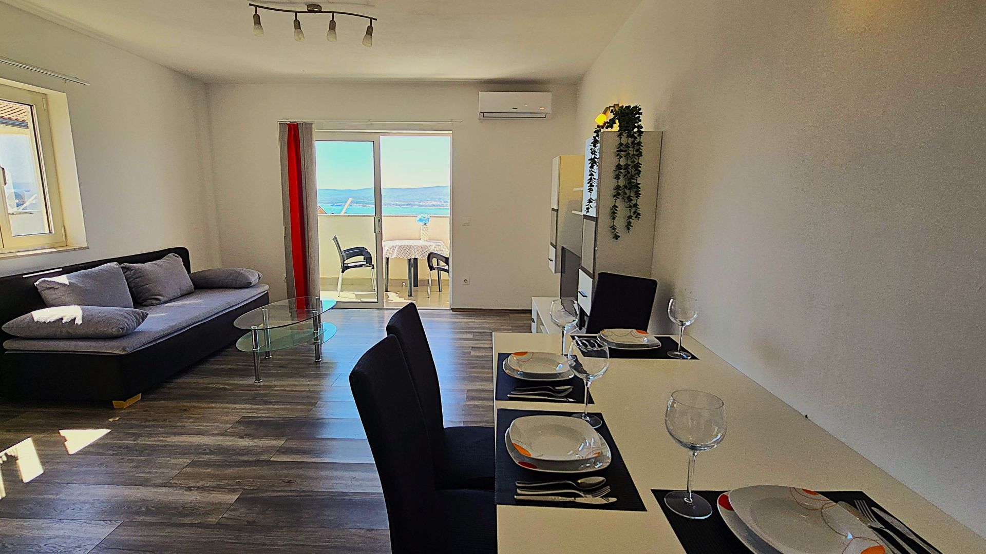 A modern living room with a sofa, a dining table set for two, and a view through a glass door to an outdoor balcony.