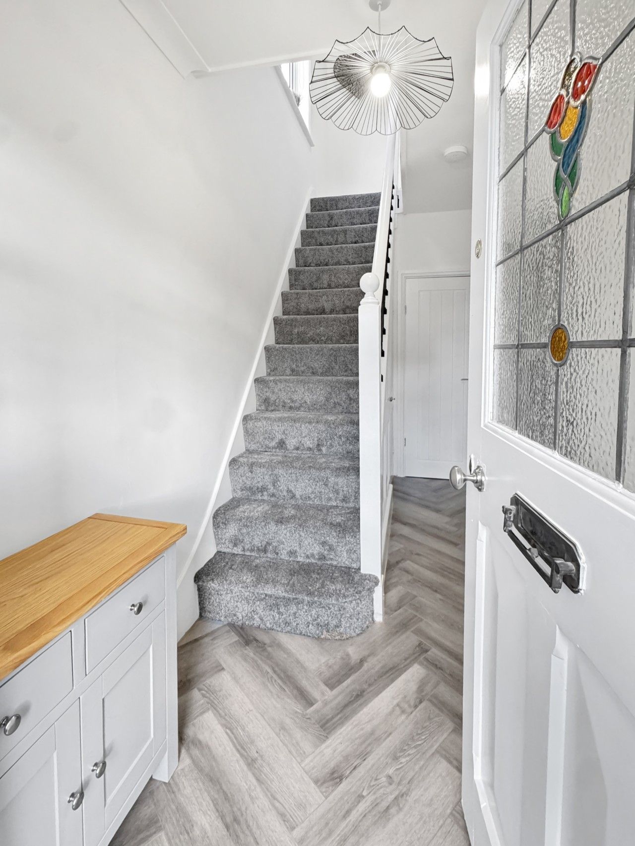 Un escalier recouvert d’une moquette grise, un banc en bois et une porte blanche avec un panneau de verre.