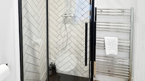 Um chuveiro com paredes de vidro e padrão geométrico de herringbone em azulejos, piso de madeira e um porta-toalhas com uma toalha branca.
