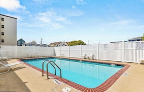 Une piscine rectangulaire entourée d’une clôture blanche, avec un poste de surveillance pour les sauveteurs et des chaises longues.