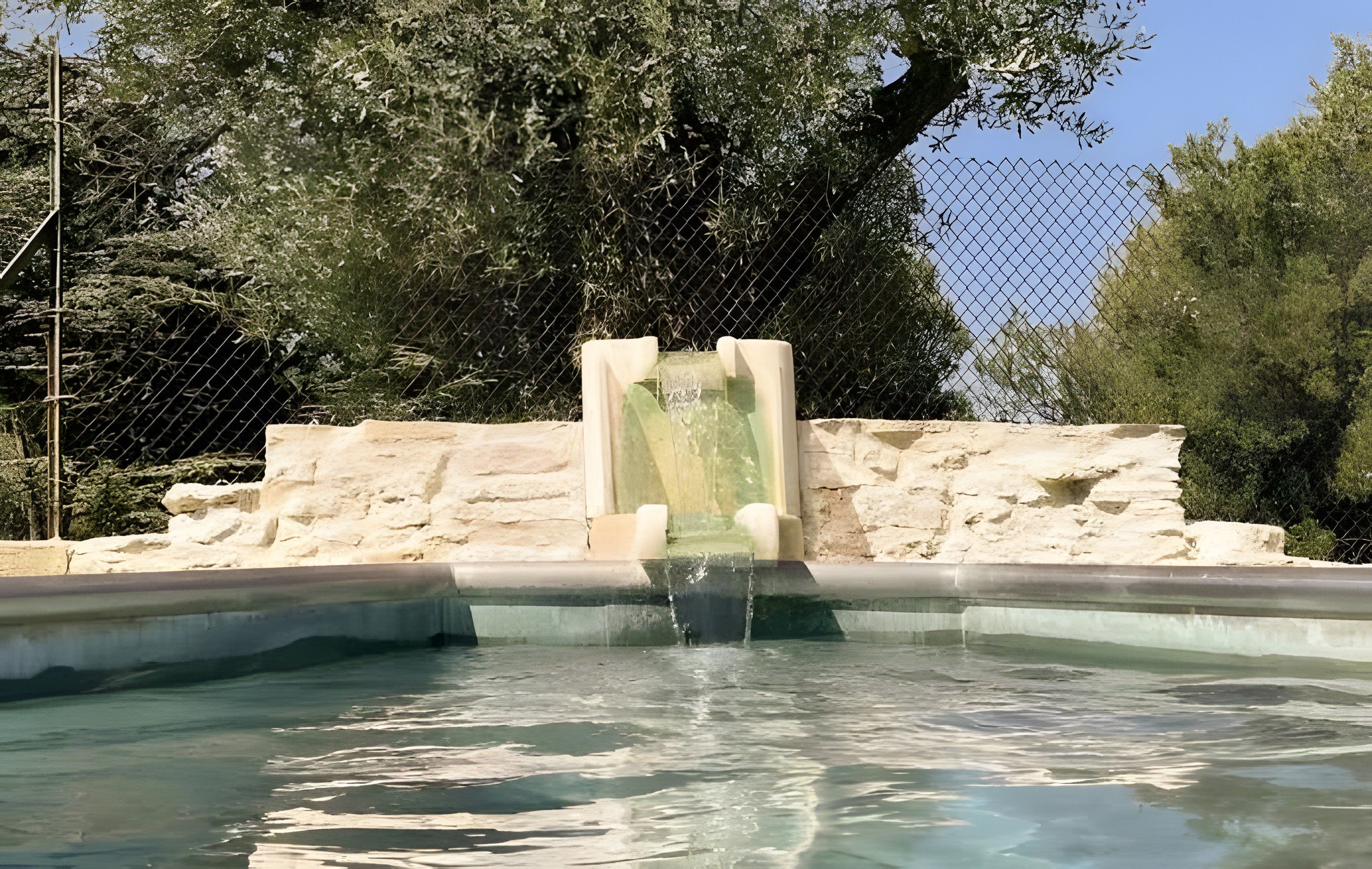 A pool with a waterfall feature, surrounded by a stone wall and a chain-link fence.