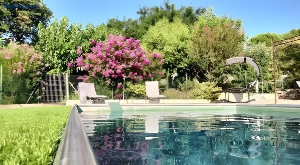 A swimming pool with lounge chairs, a covered seating area, and lush greenery in the background.