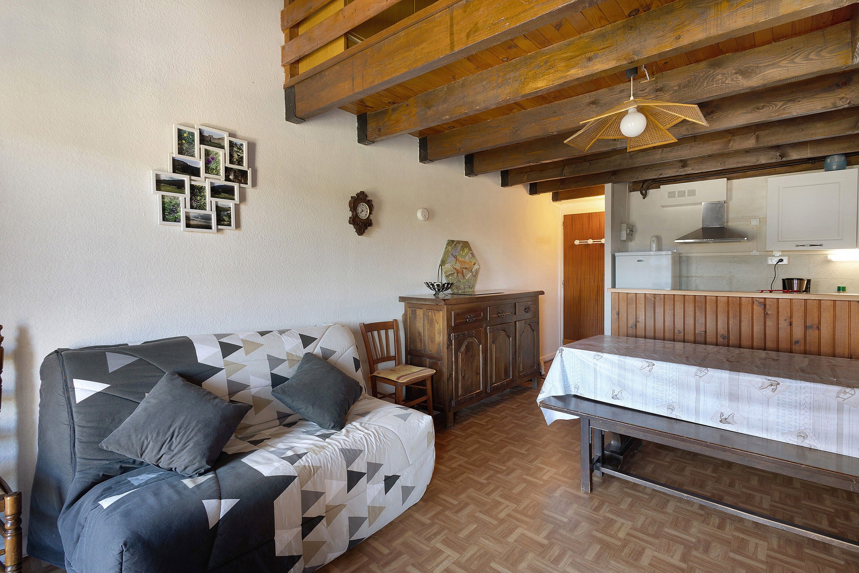 A cozy room with a wooden ceiling, a sofa with patterned cushions, a dining table, and a kitchen area with a stove.