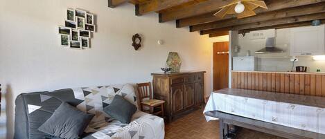 A cozy room with a wooden ceiling, a sofa with patterned cushions, a dining table, and a kitchen area with a stove.