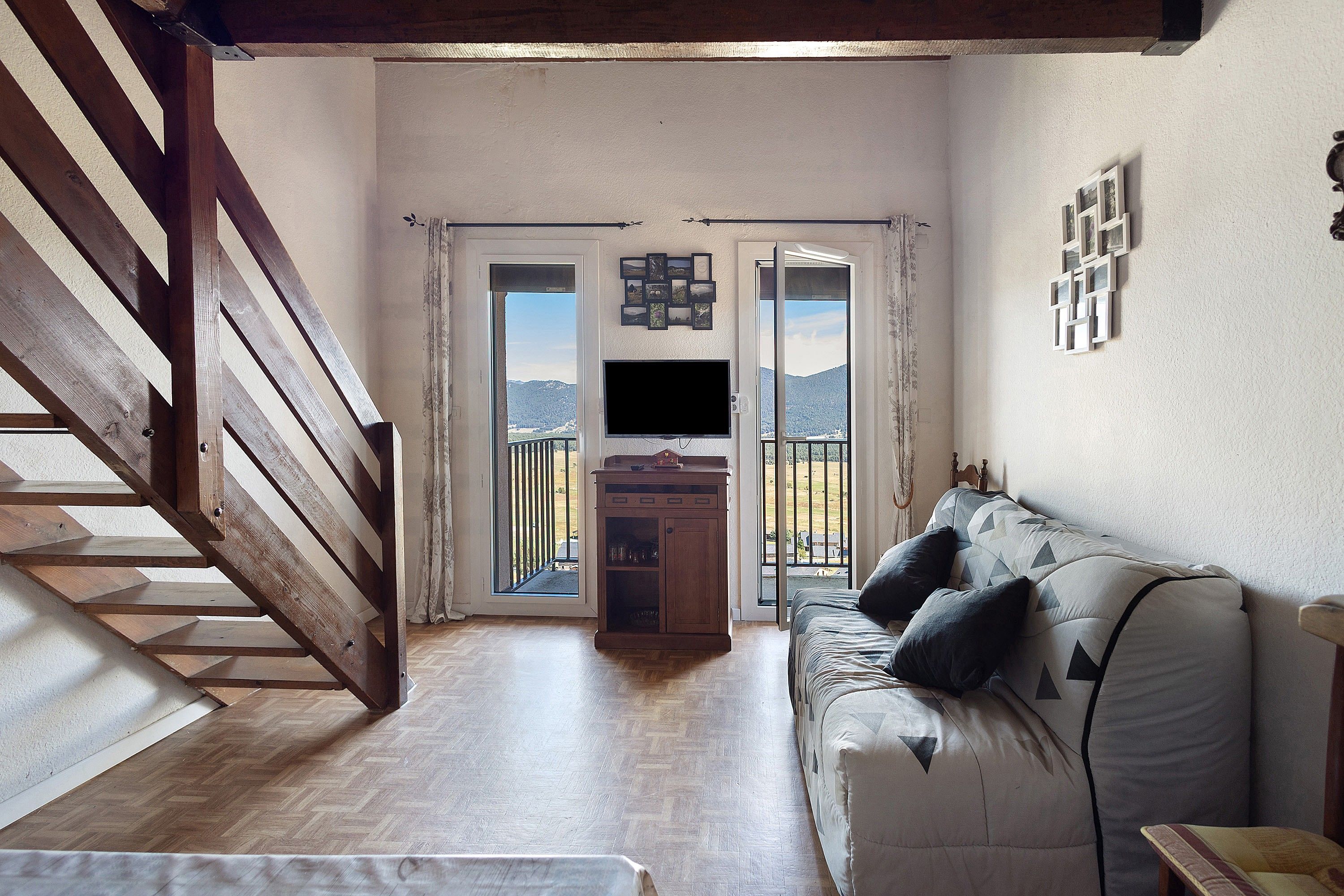 Habitació amb una escala de fusta, un sofà amb coixins, una televisió sobre un suport i una vista per una porta de vidre.