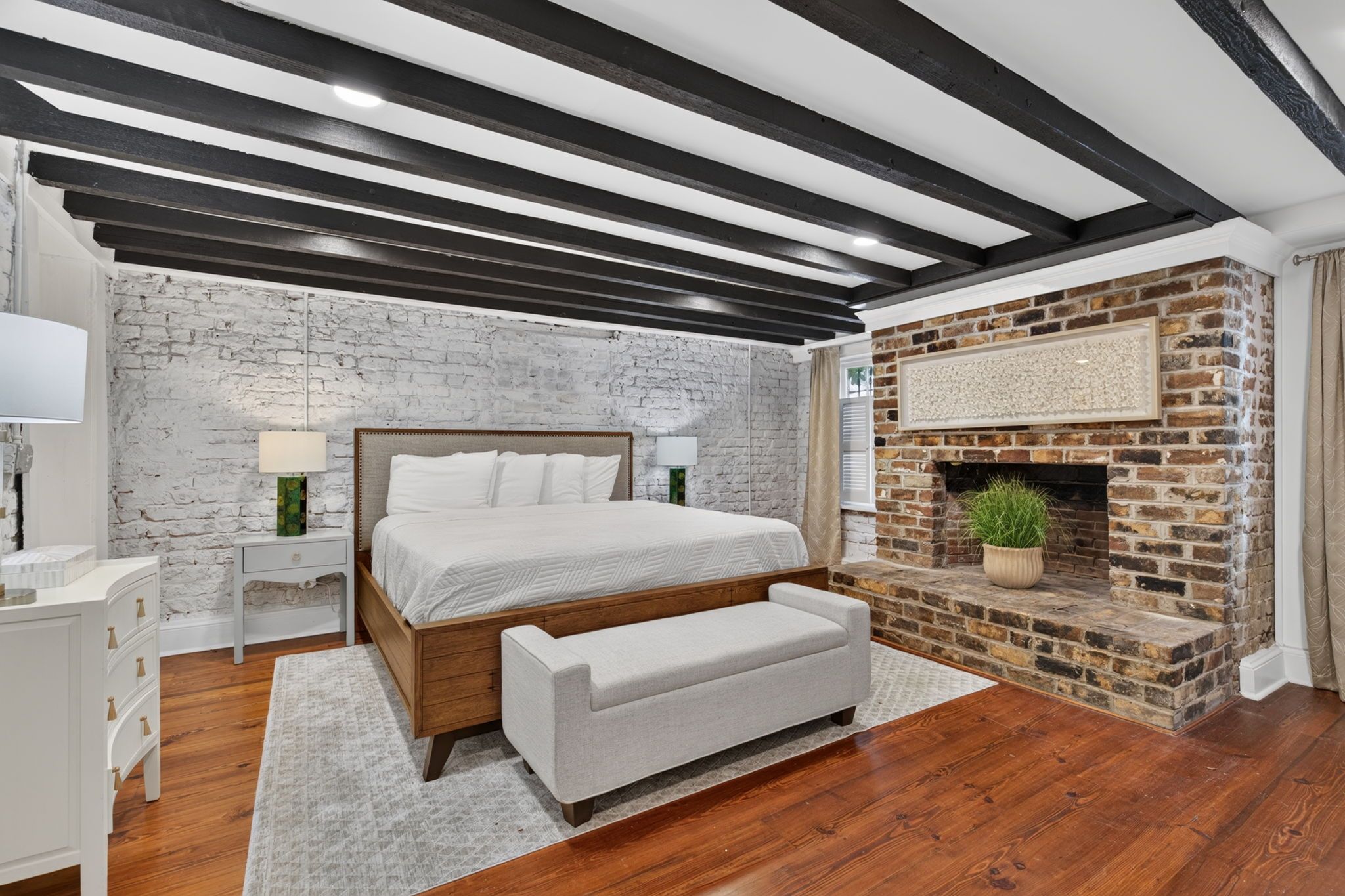 A bedroom with a bed, bedside tables, a bench, a fireplace, and exposed wooden beams on the ceiling.