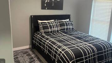 Een slaapkamer met een bed, een hoofdeinde, een aan de muur hangend kunstwerk en een raam met jaloezieën.
