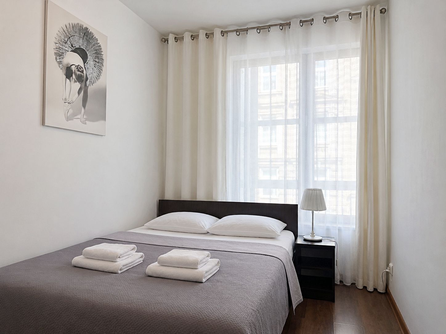 Una cama bien hecha con sábanas blancas, una lámpara de noche y una ventana con cortinas traslúcidas.