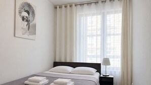 Cama bem arrumada com roupa branca, luminária de cabeceira e janela com cortinas leves.