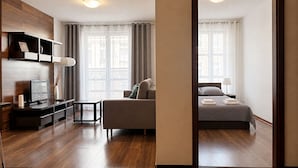 Quarto de hotel moderno com piso de madeira, sofá, cama, escrivaninha e televisão.