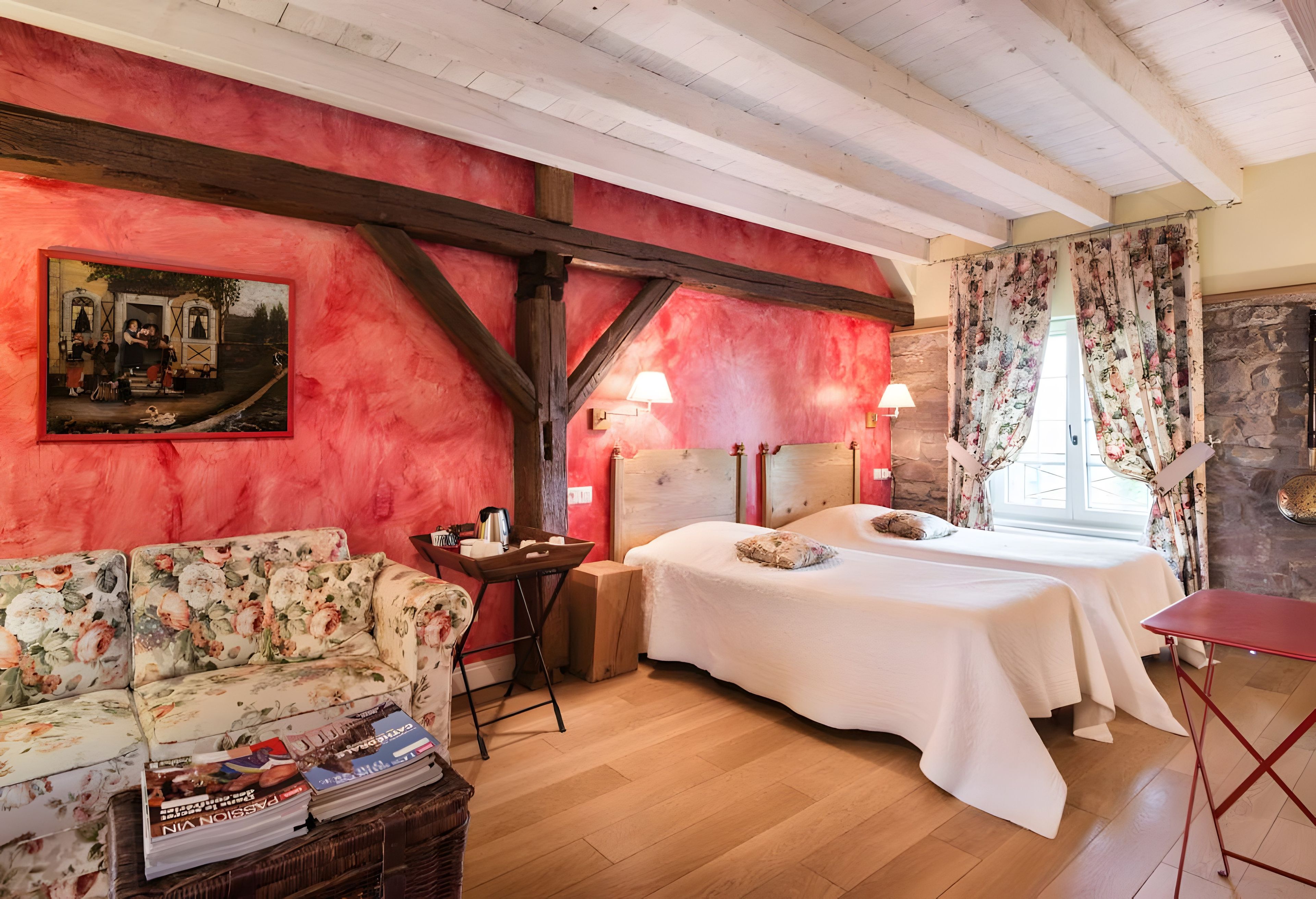 Een kamer met een houten plafond, een rode muur met een schilderij, een bloemenbank, een bed met een witte spreidek en een kleine tafel met boeken en een lamp.