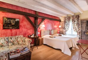 A room with a wooden ceiling, a red wall with a painting, a floral sofa, a bed with a white bedspread, and a small table with books and a lamp.