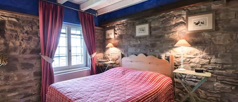 A bedroom with a stone wall, a wooden ceiling, a bed with a red checkered bedspread, two bedside lamps, and a window with curtains.