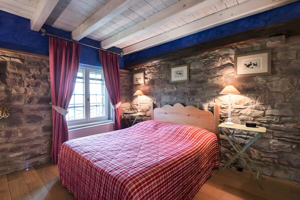 Quarto com parede de pedra, teto de madeira, cama com colcha xadrez vermelha, duas luminárias de cabeceira e janela com cortinas.