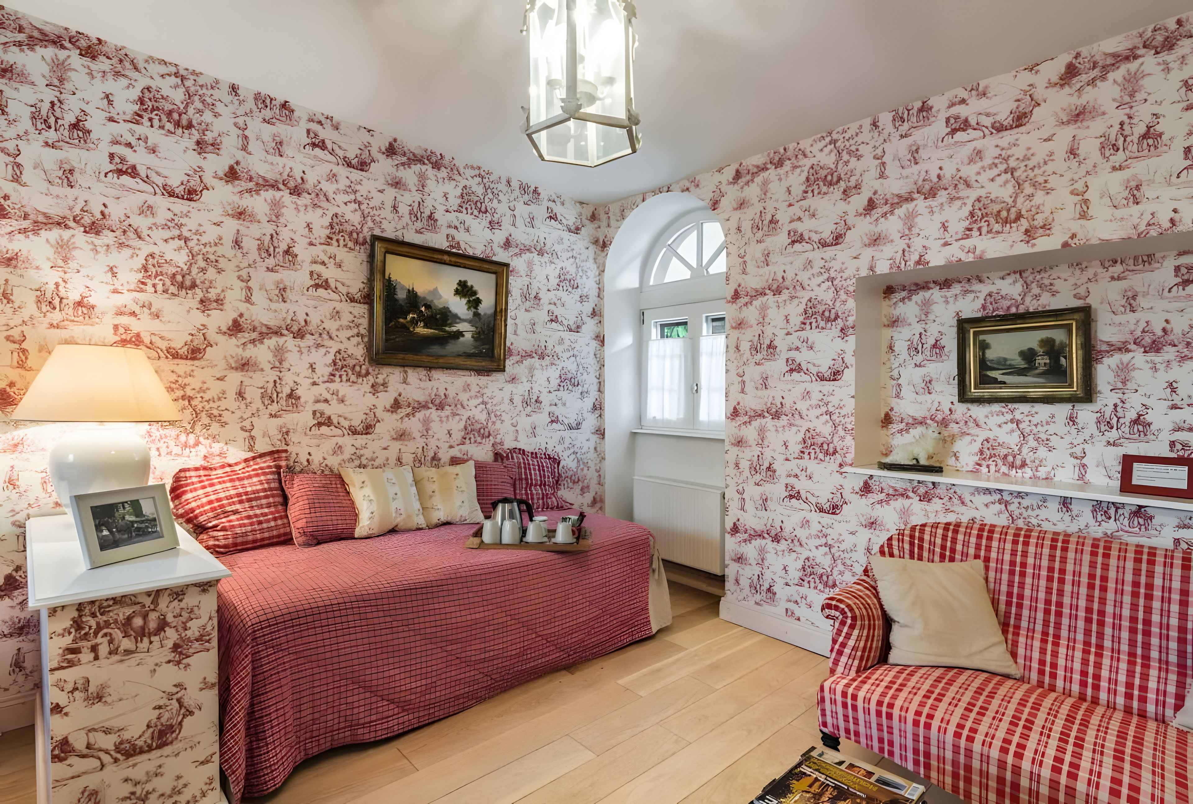 Een kamer met bloemenbehang, een bed met een rood geruit beddegoed, een witte nachtkastje met een lamp en een rode en witte geruite bank.