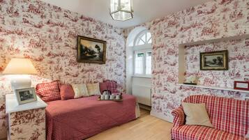 Een kamer met bloemenbehang, een bed met een rood geruit beddegoed, een witte nachtkastje met een lamp en een rode en witte geruite bank.