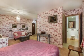 Quarto com papel de parede floral, cama, sofá, mesa pequena e porta que dá para outro cômodo.