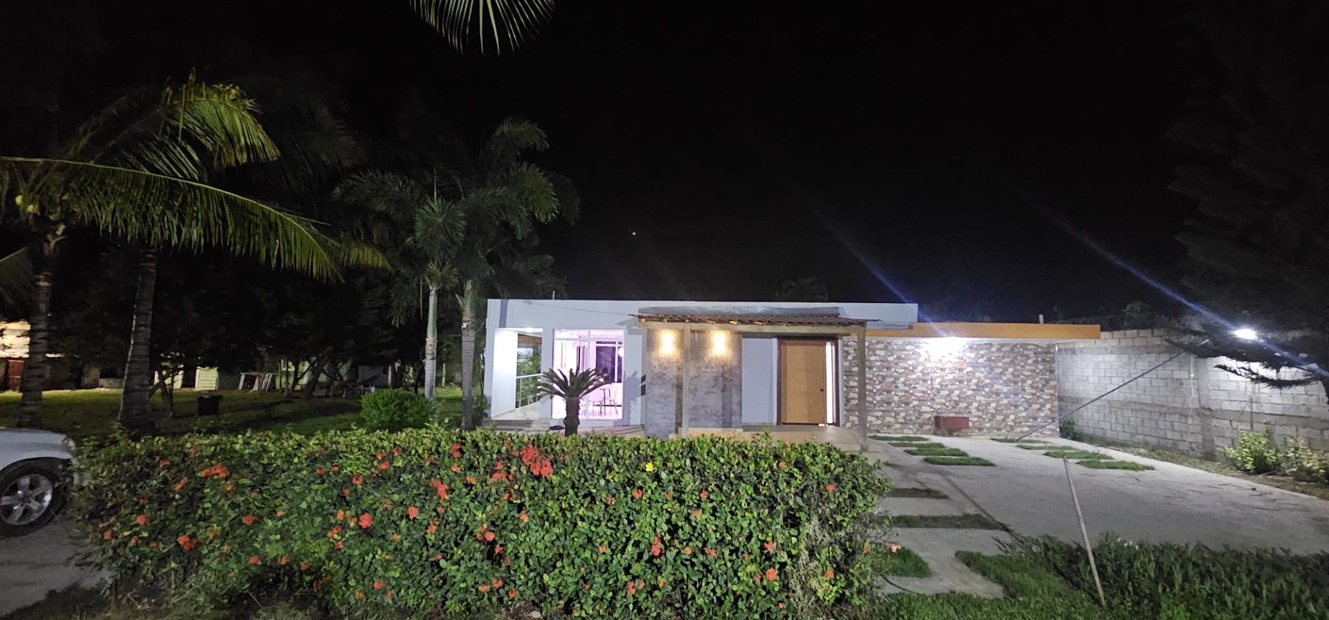Una casa moderna con una entrada iluminada, un muro de piedra y un jardín con flores rojas.